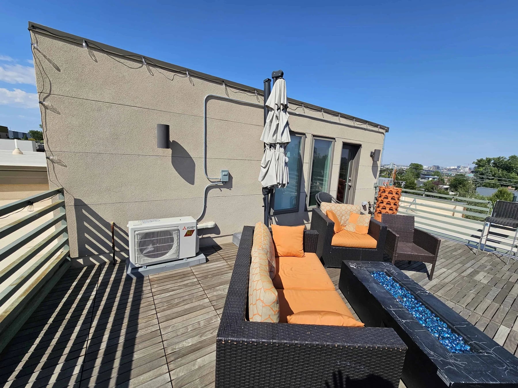 Mitsubishi single-zone outdoor unit on a rooftop deck beside orange patio cushions and a fire table, with its line set and disconnect on the wall