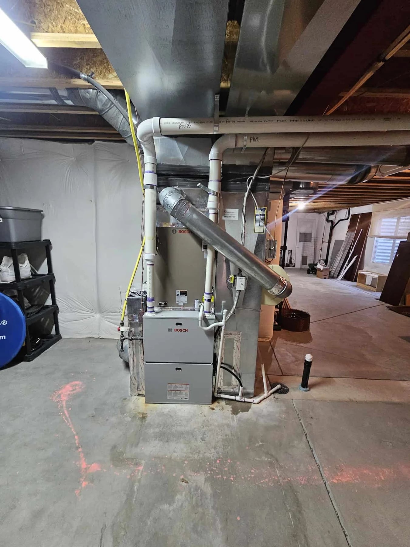 Bosch furnace in an unfinished basement with white PVC intake and exhaust pipes, a yellow gas line and a drain line to the floor