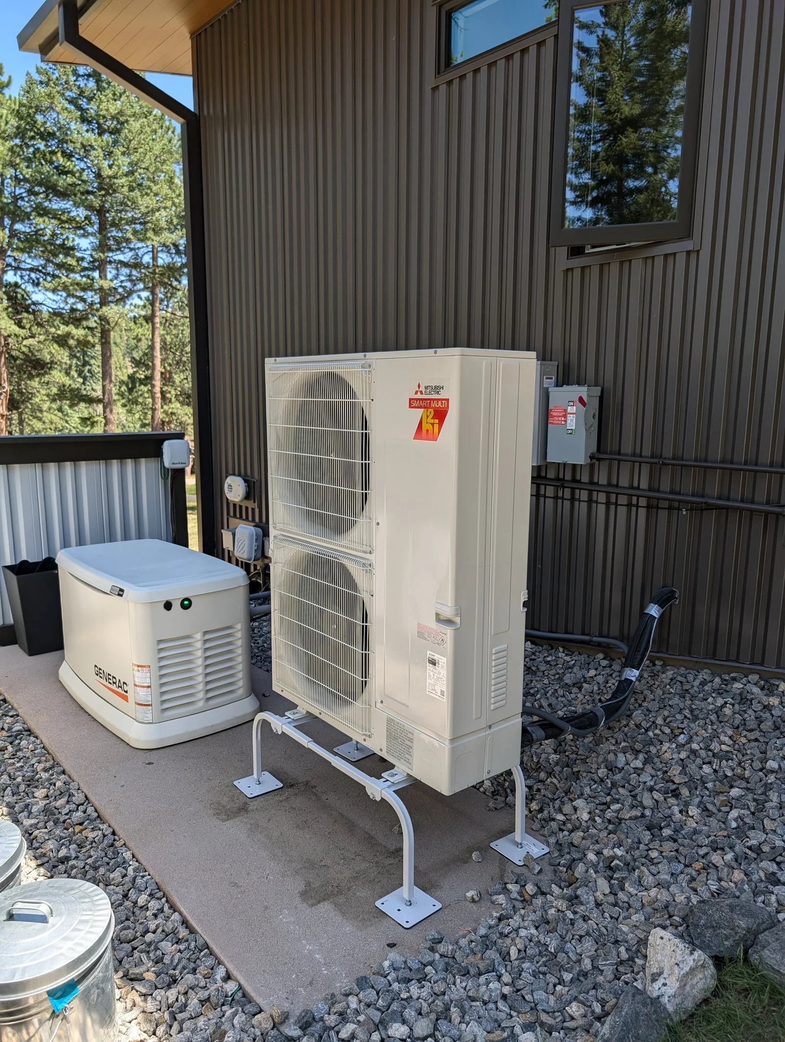A Mitsubishi two-fan heat pump on a stand beside a standby generator at a dark-sided house in the pines