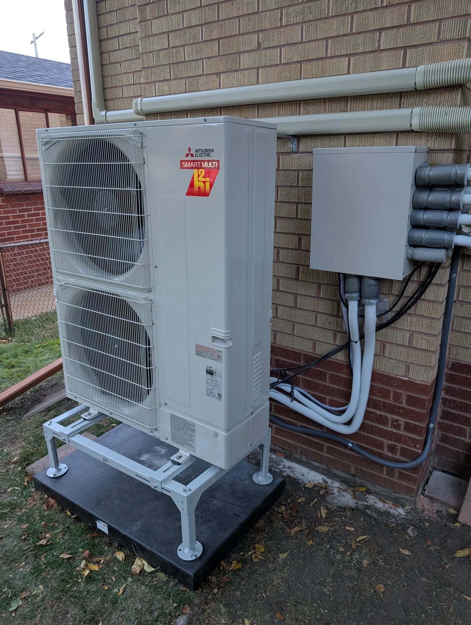 A Mitsubishi multi-zone outdoor unit on a stand beside tan brick, with a branch box on the wall