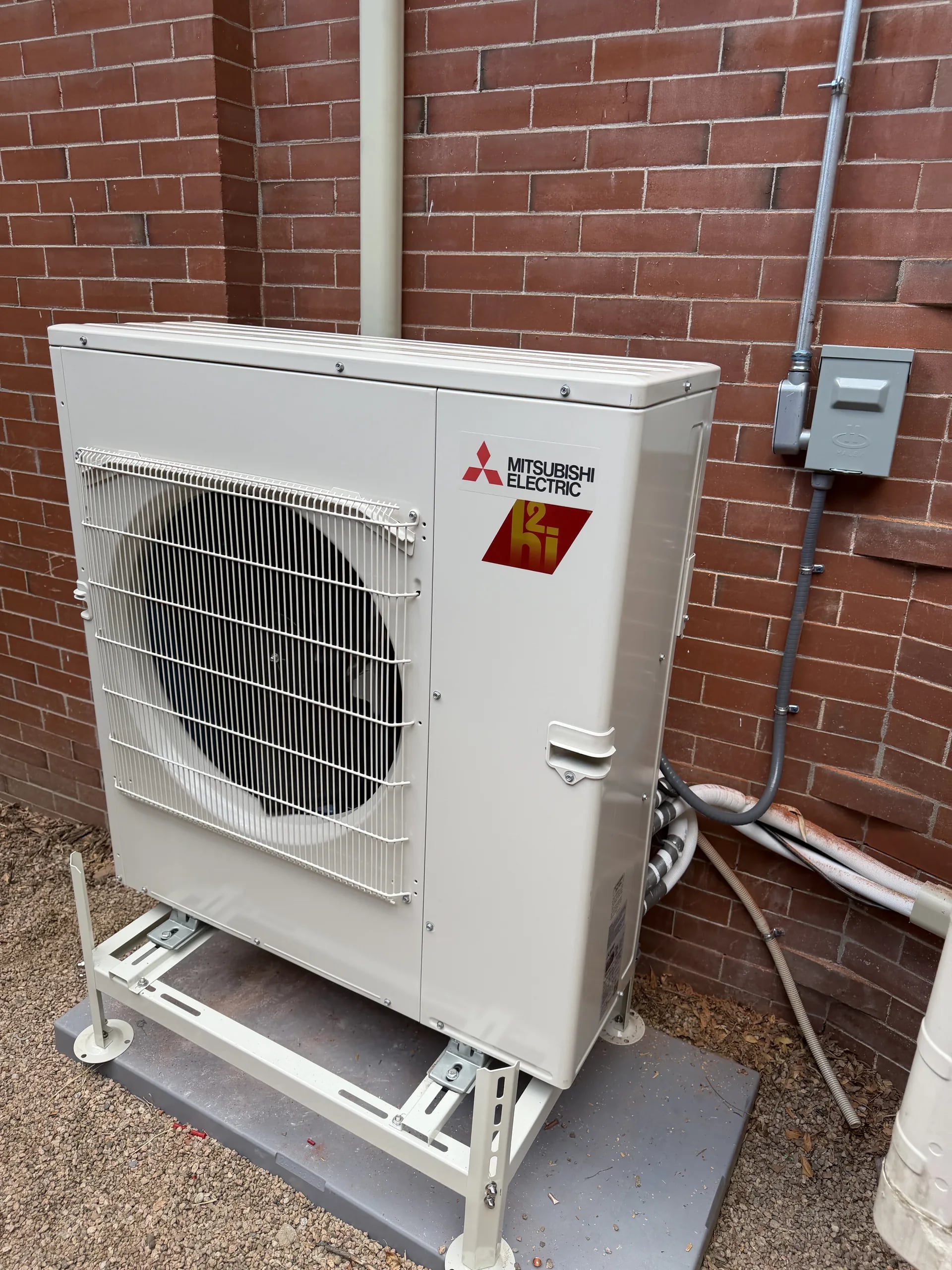 Mitsubishi two-fan outdoor unit on a stand beside red brick, with its line set in a white cover and a disconnect on the wall