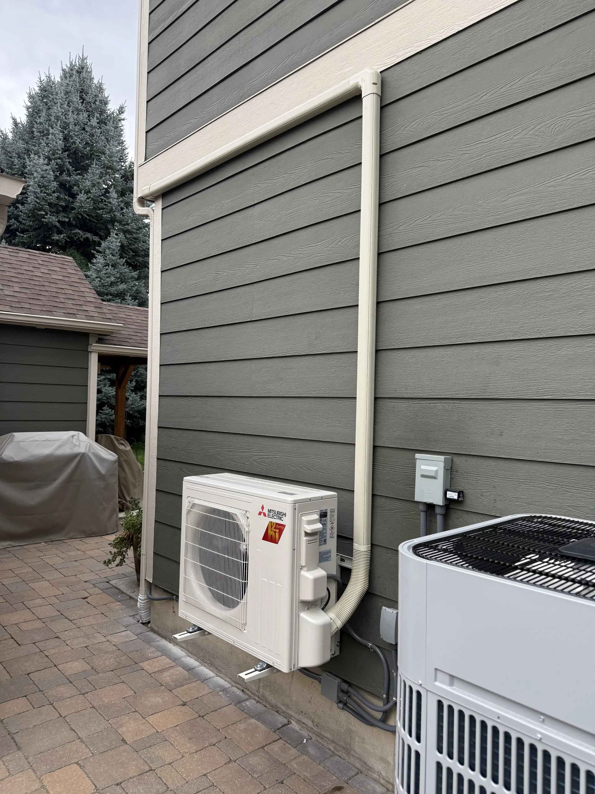 Mitsubishi outdoor unit on a stand on brick pavers beside green siding, with a line-set cover up the wall and a second condenser beside it