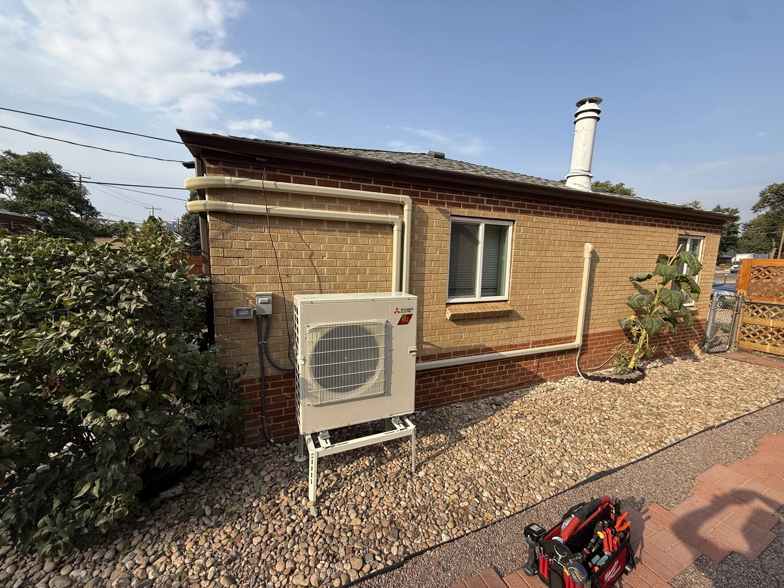 A newly installed Mitsubishi heat pump on a stand beside a tan brick ranch house, its line set covered along the wall
